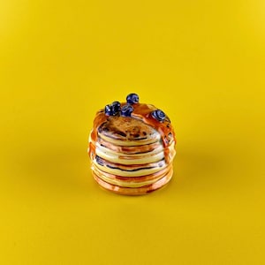 May include: A stack of pancakes with blueberries and syrup on a yellow background. The pancakes are a light brown color, with darker brown edges. The syrup is a glossy, amber color, and the blueberries are a deep blue.