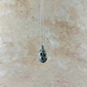 May include: A handcrafted pendant necklace featuring a teardrop-shaped ceramic pendant. The pendant is a blend of teal and brown with leaf-like cutouts, suspended from a delicate silver chain. The necklace is displayed against a neutral background.