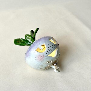 May include: A ceramic Halloween-themed turnip with a carved face and a green leafy top. The turnip is painted in shades of gray, purple, and pink, with small, textured bumps. The carved face has glowing yellow light.