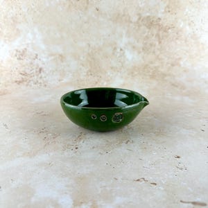 May include: A small, handcrafted, dark green ceramic bowl with a spout. The bowl has a glossy finish and three small decorative holes on the side. The bowl is designed for serving sauces or dips.