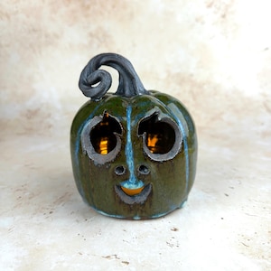 May include: A ceramic jack-o'-lantern pumpkin figurine with a glossy dark green glaze and blue highlights. The pumpkin features a carved face with two round eyes and a smiling mouth, topped with a dark, textured, spiraled stem.