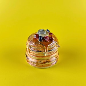 May include: A stack of ceramic pancakes with syrup dripping down the sides. The pancakes are topped with a swirl of cream, red and blue berries. The ceramic pancakes are on a yellow background.