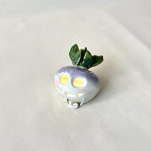 May include: A whimsical ceramic turnip figurine with a smiling face, glowing eyes, and green leafy top. The turnip is primarily white with purple and pink accents. The design has a playful, cartoonish aesthetic.
