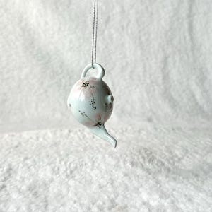 May include: A white ceramic teapot ornament with a pink floral design. The teapot has a small spout and a handle. It is hanging from a silver string.