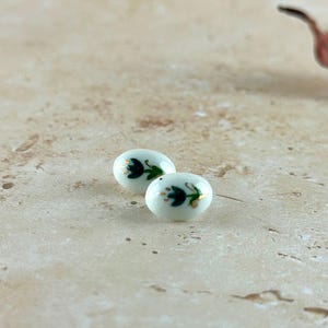 May include: Two small, oval-shaped, white ceramic beads. Each bead features a dark green floral design with gold accents. The beads are displayed on a light beige surface.