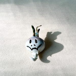 May include: A white ceramic onion with a green stem and a hand-painted sad face. The onion is sitting on a white fabric surface.