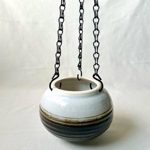 May include: A white ceramic hanging planter with brown stripes. The planter is suspended from three black chains.