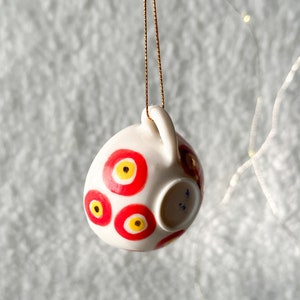 May include: White ceramic ornament with a red and yellow eye design. The ornament is hanging from a gold string.