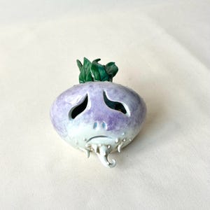 May include: A ceramic turnip-shaped decorative object with a sad face carved into the front. The turnip is primarily lavender with a white base and green leaves on top. The object has a whimsical, handcrafted appearance.