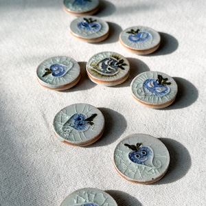 May include: Nine ceramic buttons with a white background and blue and brown floral designs. The buttons have a raised heart shape in the center.