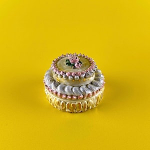 May include: A two-tiered ceramic cake-shaped trinket box. The box is yellow with white, pink, and gold accents. The top tier is decorated with pink roses. The box is set against a yellow background.