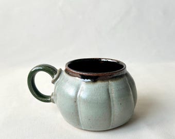 May include: A small, light green ceramic mug with a rounded body and a dark green handle. The rim is brown, and the interior is black. The mug has vertical ridges and a pumpkin-like shape, suggesting a seasonal or rustic theme.