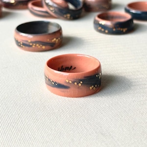 May include: A collection of ceramic rings with a wide band design. The rings feature a coral-colored base with a dark blue band and gold speckles. Some rings have a handwritten inscription. The rings are displayed on a white surface.