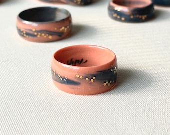 May include: A collection of ceramic rings with a wide band design. The rings feature a coral-colored base with a dark blue band and gold speckles. Some rings have a handwritten inscription. The rings are displayed on a white surface.