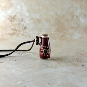May include: A small, red ceramic pitcher pendant with a cork stopper and a brown leather cord. The pitcher features a white rim and a gold-centered flower design. The pendant is a unique piece of jewelry.
