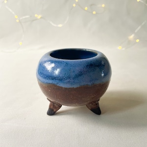 May include: A small, blue and brown ceramic pot with three legs. The pot is glazed in a light blue color with a brown rim and base.