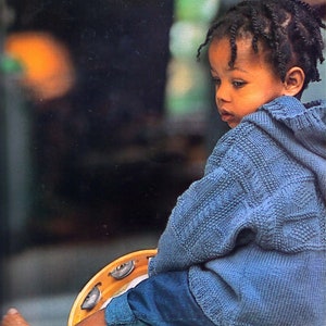 May include: A young child with braided hair wearing a blue knitted sweater sits on a wooden surface holding a tambourine.