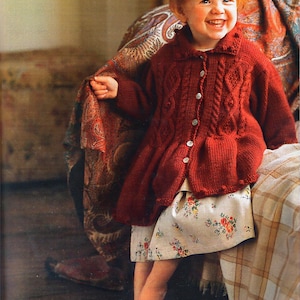 May include: A young girl with red hair wearing a red knitted cardigan with a cable knit pattern and a white floral skirt. She is sitting on a couch with a patterned throw blanket.