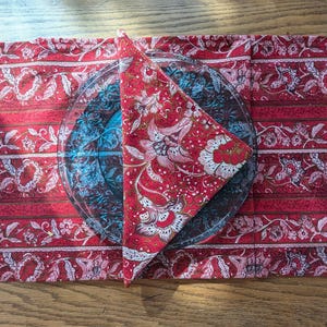 May include: A red and white patterned fabric placemat with a red and white patterned fabric napkin folded into a triangle on top of it. The napkin is on top of a clear glass plate.
