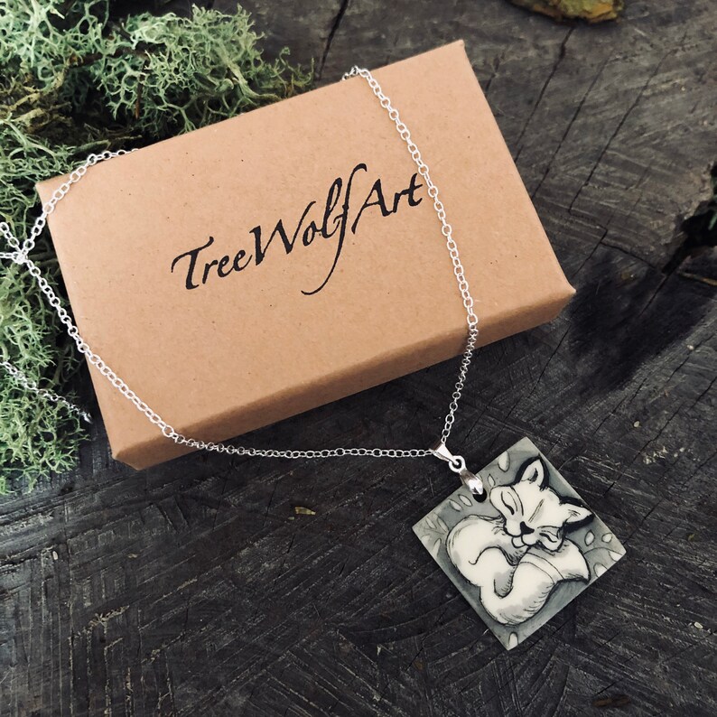 May include: A silver chain necklace with a square pendant depicting a sleeping fox. The pendant is grey and white, and the necklace is presented on a wooden surface. The necklace is displayed with a brown box that has the text "TreeWolfArt".