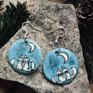 May include: Circular blue earrings with a charming design. Each earring displays a crescent moon and two white foxes looking upwards, set against a starry sky. The earrings have silver-coloured hooks.