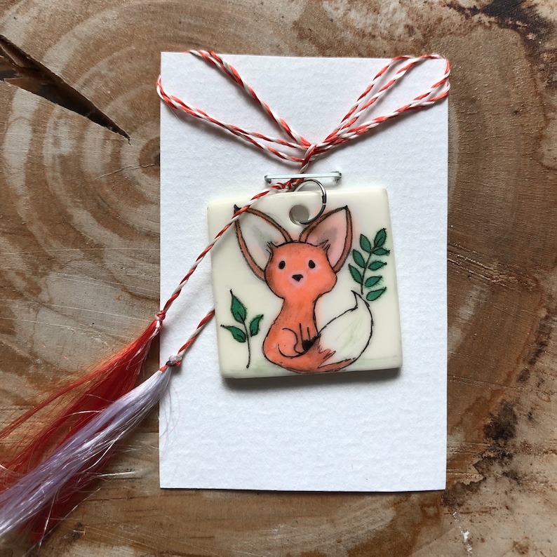 May include: A square pendant with a cartoon fox illustration in orange, white, and black, with green leaf accents. The pendant hangs from a red and white twisted cord with a tassel. The pendant is on a white card.