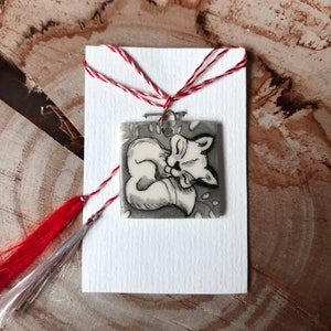May include: A square pendant with a sleeping fox illustration in black and white, attached to a white card with red and white string. The fox is curled up, with its eyes closed. The pendant is on a wooden surface.