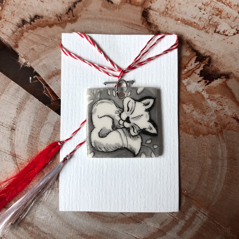 May include: A square pendant with a sleeping fox illustration in black and white, attached to a white card with red and white string. The fox is curled up, with its eyes closed. The pendant is on a wooden surface.