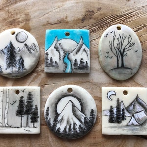 May include: Hand-painted pendants with nature-themed designs. The pendants feature mountain ranges, trees, and a camping scene, all in shades of grey and black with a touch of blue. Each pendant has a small hole for stringing.