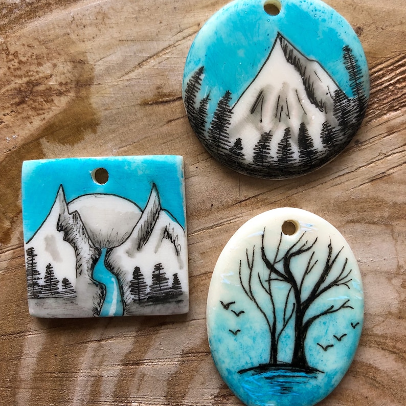May include: Three handmade pendants featuring landscape designs. One is round with a mountain scene, another is square with a mountain and river scene, and the last is oval with a tree and birds. Each has a hole for stringing.