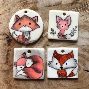 May include: Four hand-painted fox pendants. The pendants are in various shapes: round and square. Each pendant features a different fox design in shades of orange, white, and black. The pendants have small holes for hanging.