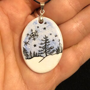 May include: Oval pendant with a winter scene. The pendant features a hand-painted design of a snowy landscape with black trees, a snowflake, and stars against a blue sky. It hangs from a black cord with a silver-colored bail.