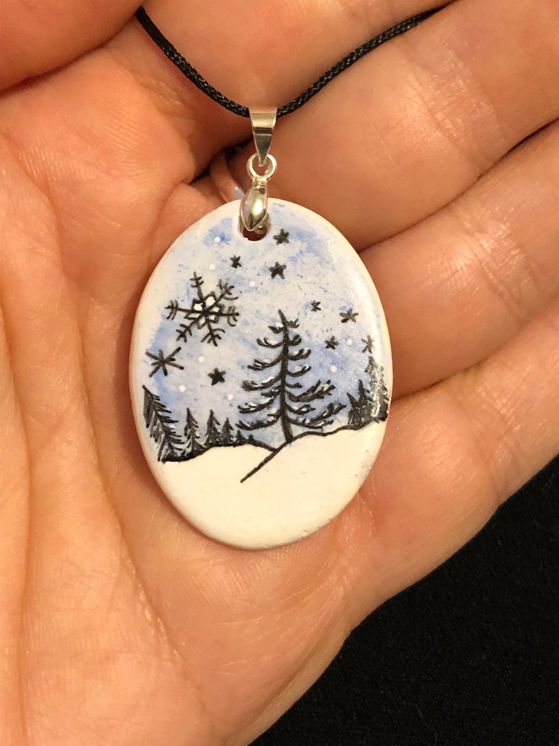 May include: Oval pendant with a winter scene. The pendant features a hand-painted design of a snowy landscape with black trees, a snowflake, and stars against a blue sky. It hangs from a black cord with a silver-colored bail.