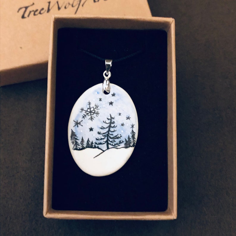 May include: Oval pendant necklace featuring a winter landscape scene. The pendant has a white base with a hand-painted design of a snowy scene with a snowflake, trees, and stars. The pendant is on a black cord and presented in a gift box.