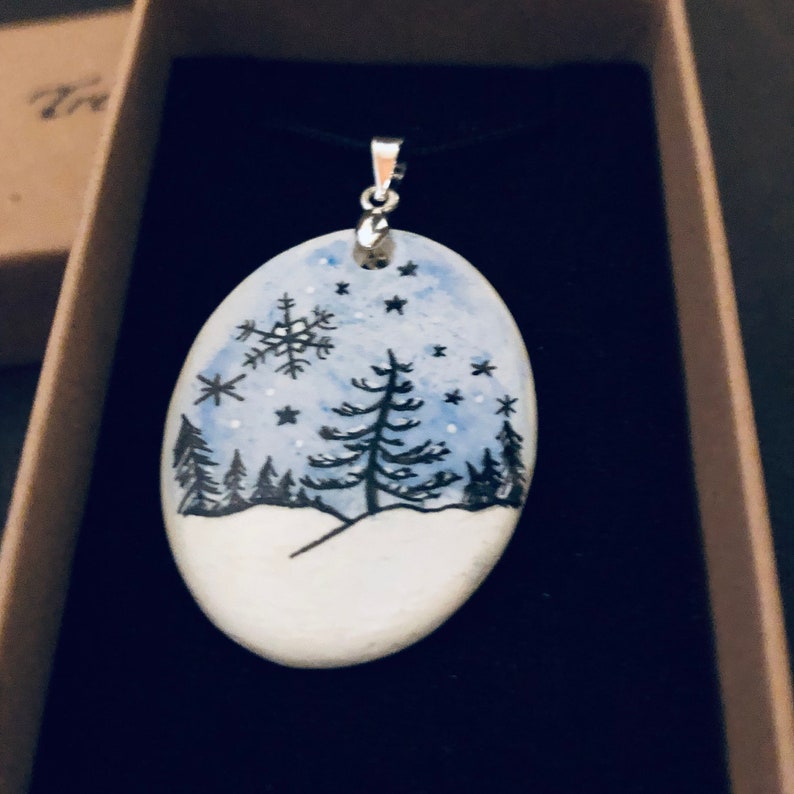 May include: Oval pendant with a winter scene. The pendant features a hand-painted design of a snowy landscape with pine trees, snowflakes, and stars against a blue sky. It hangs from a black cord with a silver-colored bail.
