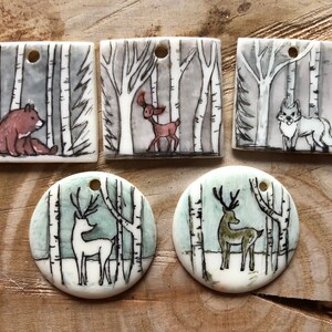 May include: Hand-painted ceramic pendants with forest scenes. The pendants depict a bear, deer, and fox in a birch forest setting. The pendants are square and round with a hole for stringing.