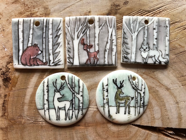 May include: Hand-painted ceramic pendants with forest scenes. The pendants depict a bear, deer, and fox in a birch forest setting. The pendants are square and round with a hole for stringing.