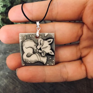 May include: A square pendant necklace with a black cord, featuring a sleeping fox illustration in black and white. The fox is curled up on a grey background with leaf details.
