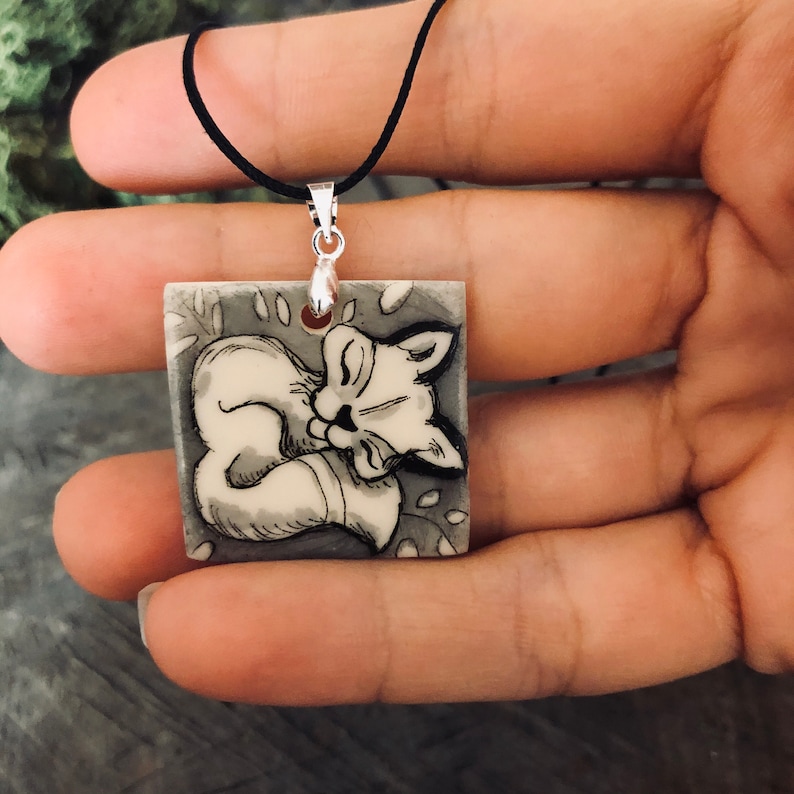 May include: A square pendant necklace with a black cord, featuring a sleeping fox illustration in black and white. The fox is curled up on a grey background with leaf details.