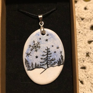 May include: Oval pendant with a winter scene. The pendant features a hand-painted design of a snowy landscape with trees, snowflakes, and stars. The pendant is on a black cord and is set in a brown box.