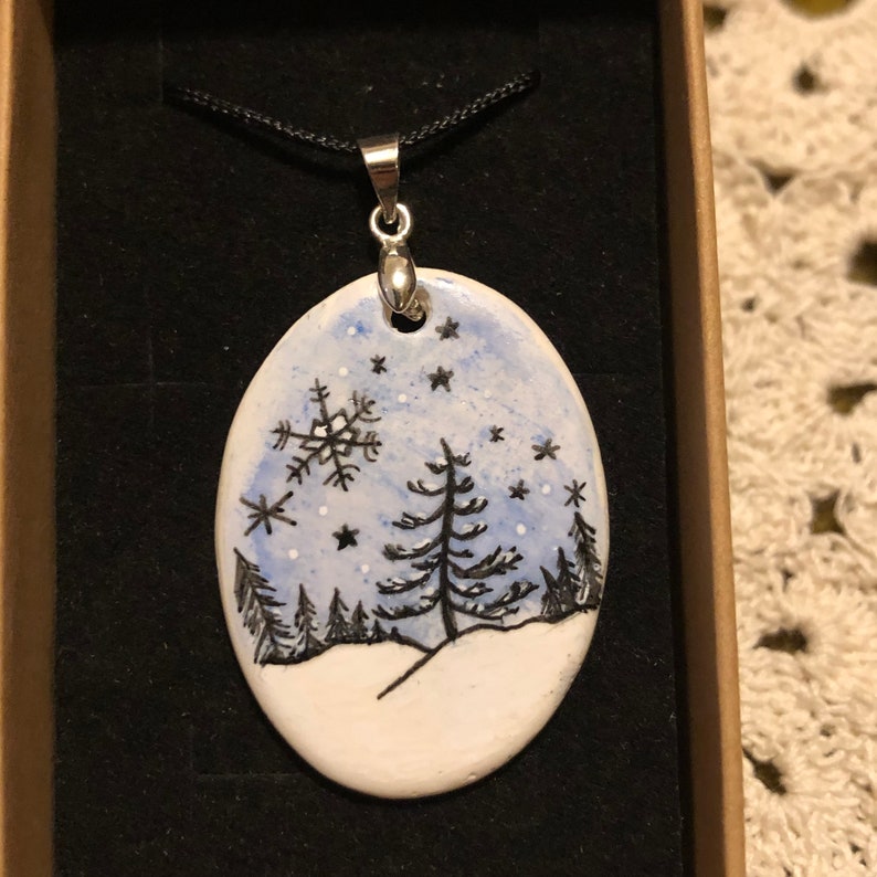 May include: Oval pendant with a winter scene. The pendant features a hand-painted design of a snowy landscape with trees, snowflakes, and stars. The pendant is on a black cord and is set in a brown box.
