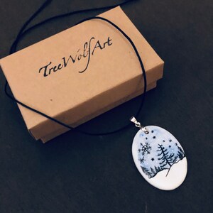 May include: A hand-painted oval pendant necklace featuring a winter landscape scene with trees, snowflakes, and stars. The pendant hangs on a black cord and rests next to a brown box with the text "TreeWolfArt".