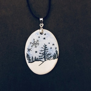 May include: Oval pendant necklace with a winter scene. The pendant features a hand-painted design of a snowy landscape with a snowflake, trees, and stars. The pendant is attached to a black cord necklace with a silver-colored bail.