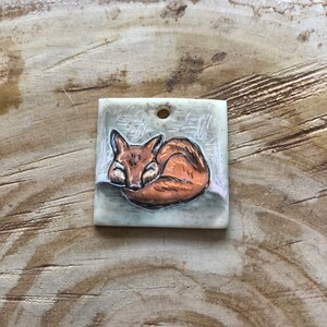 May include: Square pendant with a sleeping fox illustration. The fox is in brown and orange, curled up on a grey background with a forest design. The pendant has a small hole at the top for hanging.