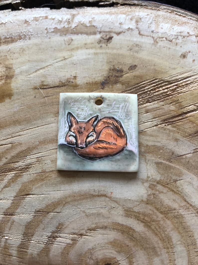 May include: Square pendant with a sleeping fox illustration. The fox is in brown and orange, curled up on a grey background with a forest design. The pendant has a small hole at the top for hanging.