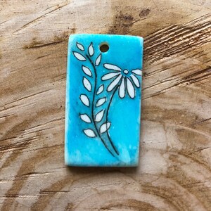 May include: Rectangular pendant with a turquoise background and a floral design. The design features a white daisy and a leafy stem with white leaves, all outlined in black. A small hole is at the top for hanging.