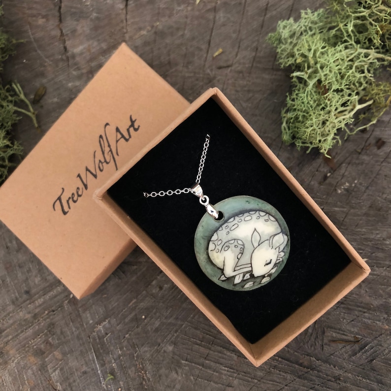May include: A silver necklace with a circular pendant depicting a sleeping fawn. The pendant is displayed in a black box with the text "TreeWolfArt" on the lid. The necklace is presented on a wooden surface.