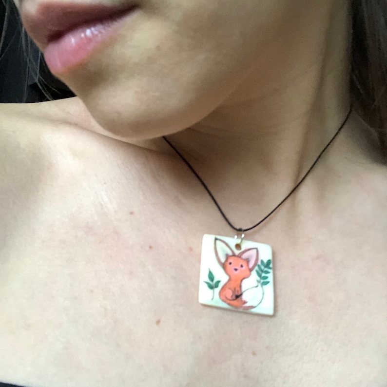 May include: A square pendant necklace with a hand-painted fox illustration. The fox is orange with a white belly and tail, surrounded by green leaves. The pendant hangs from a black cord.
