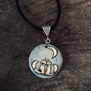 May include: A round pendant necklace featuring two foxes gazing at a crescent moon under a starry sky. The pendant is white and light blue with a black cord. The pendant is approximately 3.8 cm in diameter.