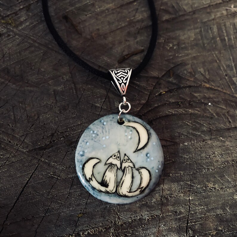 May include: A round pendant necklace featuring two foxes gazing at a crescent moon under a starry sky. The pendant is white and light blue with a black cord. The pendant is approximately 3.8 cm in diameter.
